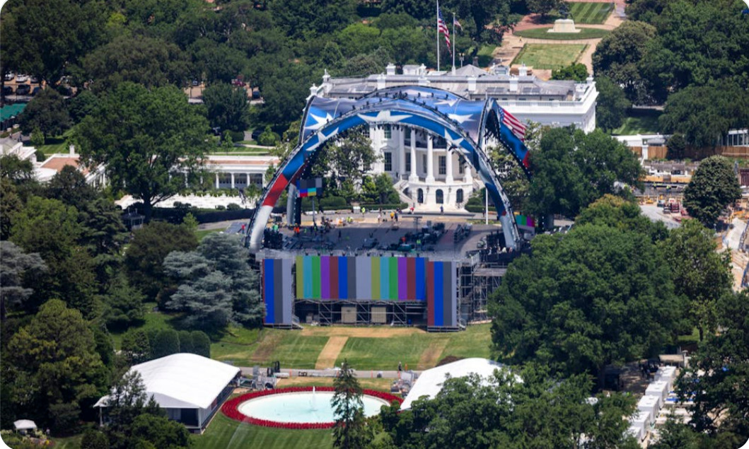Aerial view of a concert stage set up on the White House lawn, with a blue arch and colorful backdrop framed by trees and flags.