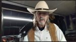 Older man in a white cowboy hat, glasses, vest, and beard speaks into a large microphone in a recording setup.