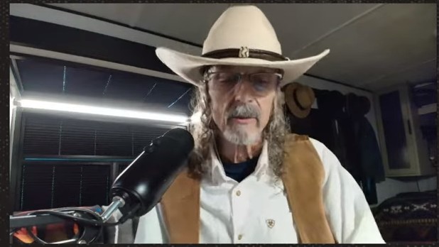 Older man in a white cowboy hat, glasses, vest, and beard speaks into a large microphone in a recording setup.