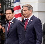 Two men in dark suits stand in front of an American flag as photographers snap nearby, looking serious and focused.