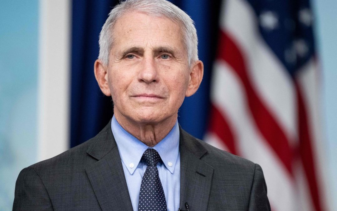 Portrait of an older man in a dark suit and blue shirt, wearing a polka-dot tie, with an American flag in the background.
